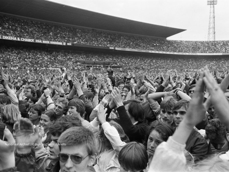 Publiek bij het Simon & Garfunkel‑concert op 12 juni 1982, gezien vanaf het podium met toeschouwers direct voor de camera en het overvolle veld en tribunes op de achtergrond.