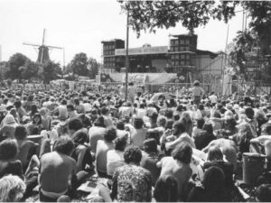 Grote menigte bezoekers voor het podium van het Holland Pop Festival 1970, met windmolen en bomen op de achtergrond.